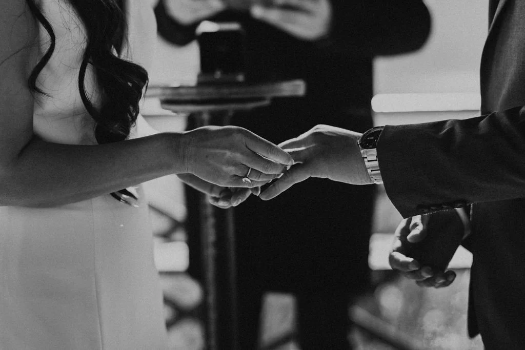 A bride and groom holding hands during their wedding ceremony. The bride is wearing a white dress and the groom is in a dark suit. They are exchanging rings or holding hands in a close-up shot.