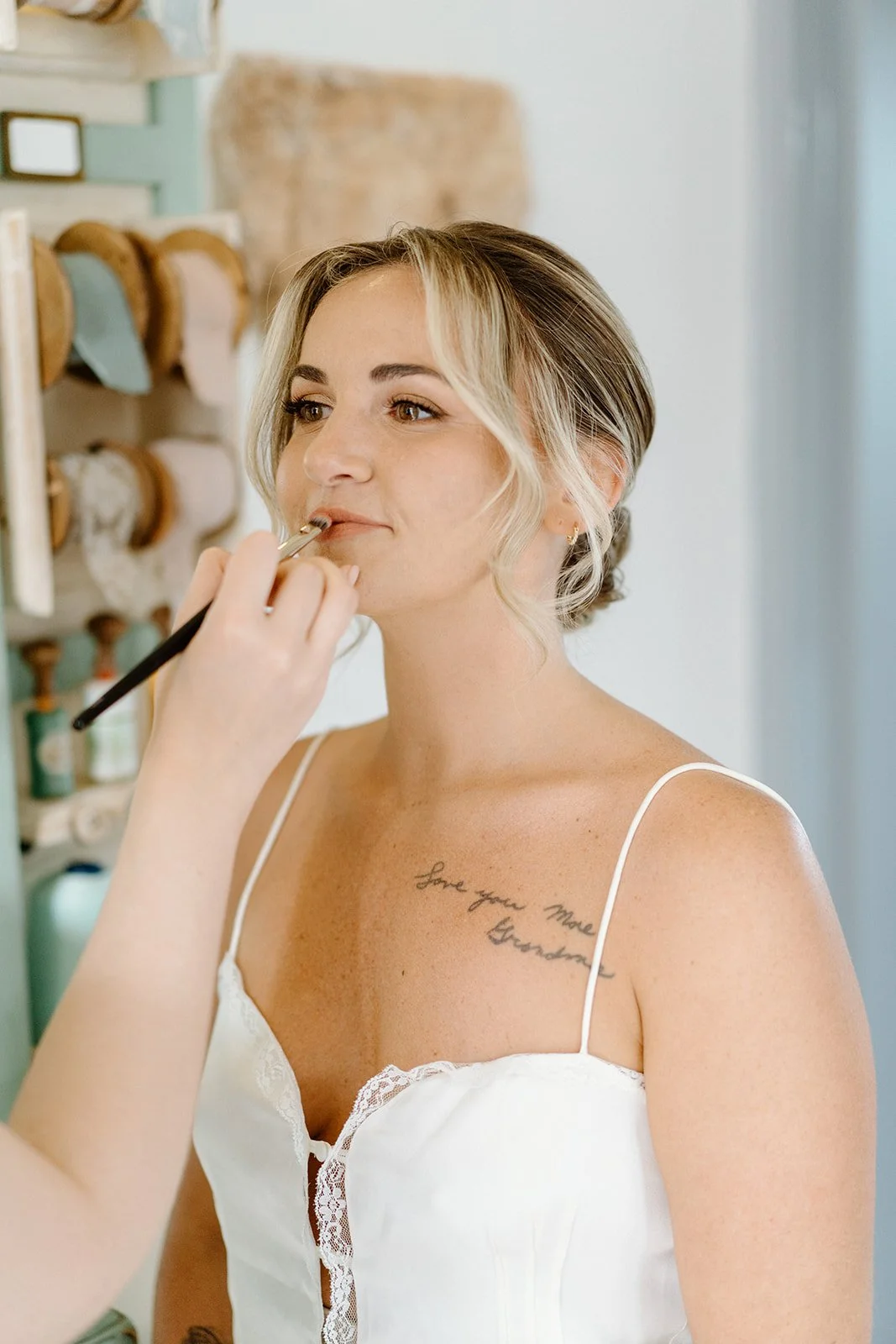 A woman getting her makeup done, wearing a white spaghetti strap top, with a visible tattoo on her chest that reads 'Love you more Grandma'.