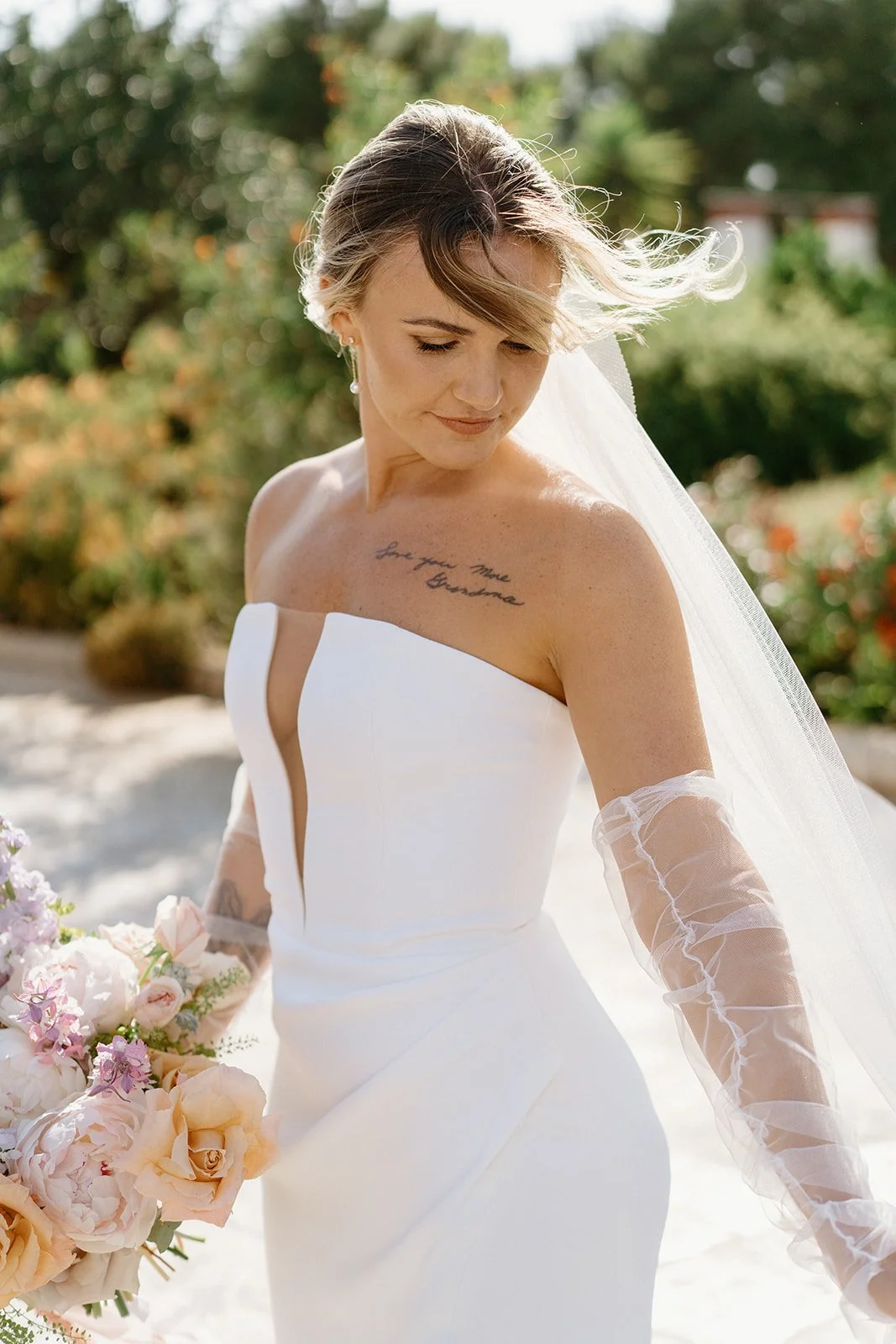 A bride in a white wedding dress holding a bouquet of flowers outdoors with greenery in the background.