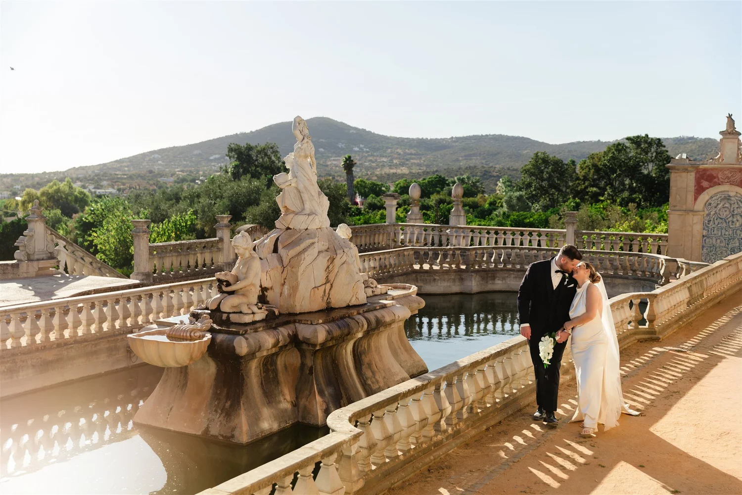 A bride and groom standing together on a stone balcony with intricate balustrades and sculptures near a large fountain. The bride wears a white satin dress and holds a bouquet, while the groom is dressed in a black tuxedo. In the background, there ar