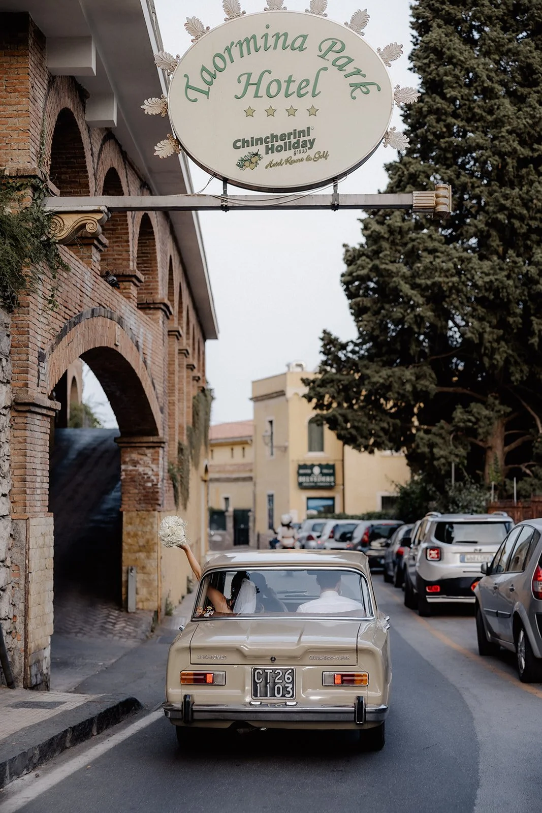 A vintage beige car driving on a narrow street with a bride and groom inside, the bride holding a bouquet of flowers, and a hotel sign overhead for the Haormina Park Hotel.