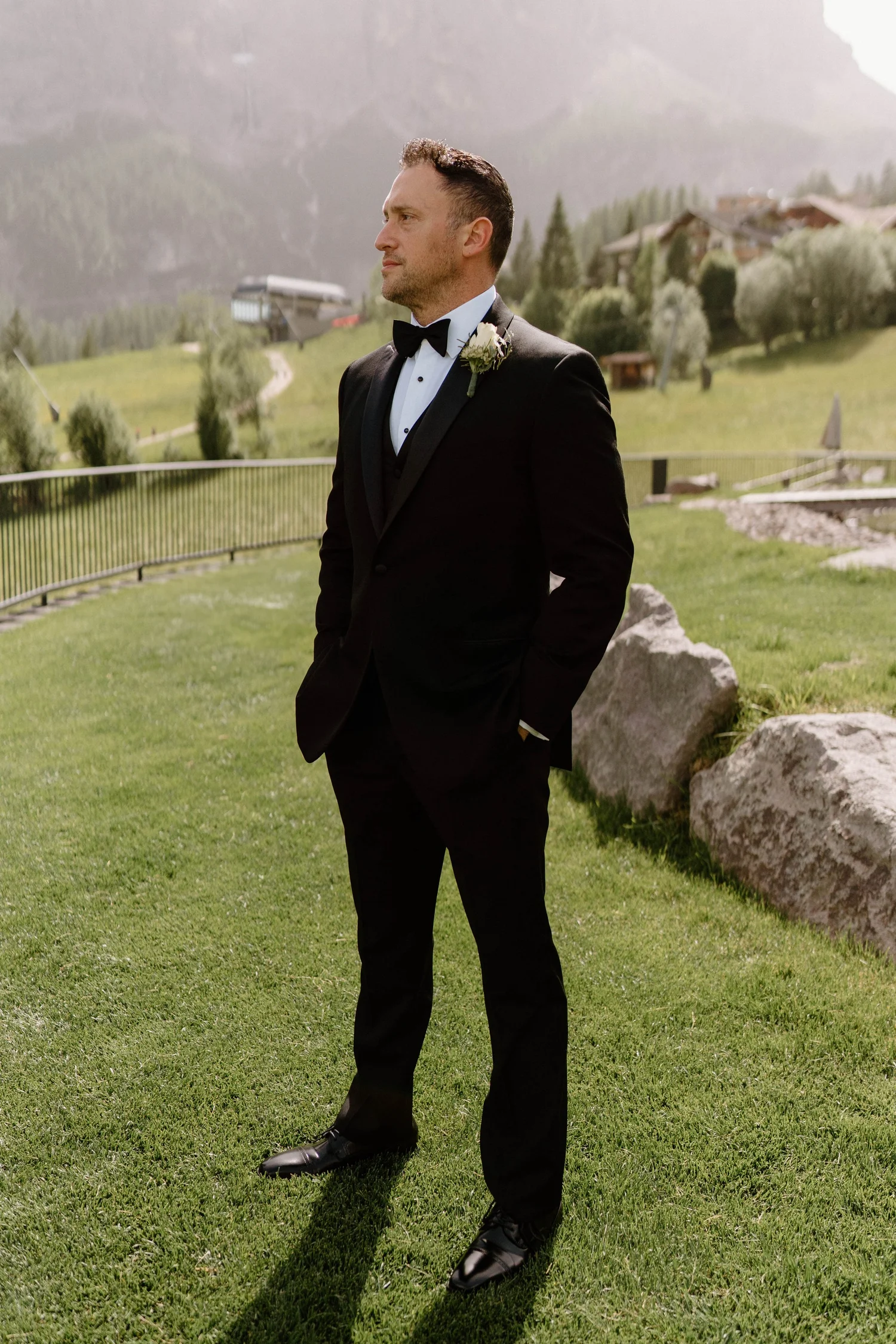 A man dressed in a black tuxedo with a bow tie, standing outdoors on grass with mountains and trees in the background.