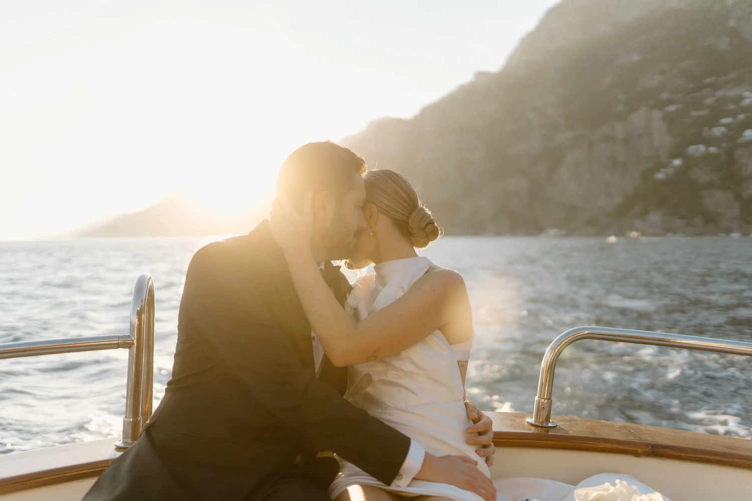 A couple sharing a kiss on a boat at sunset with a scenic island in the background.