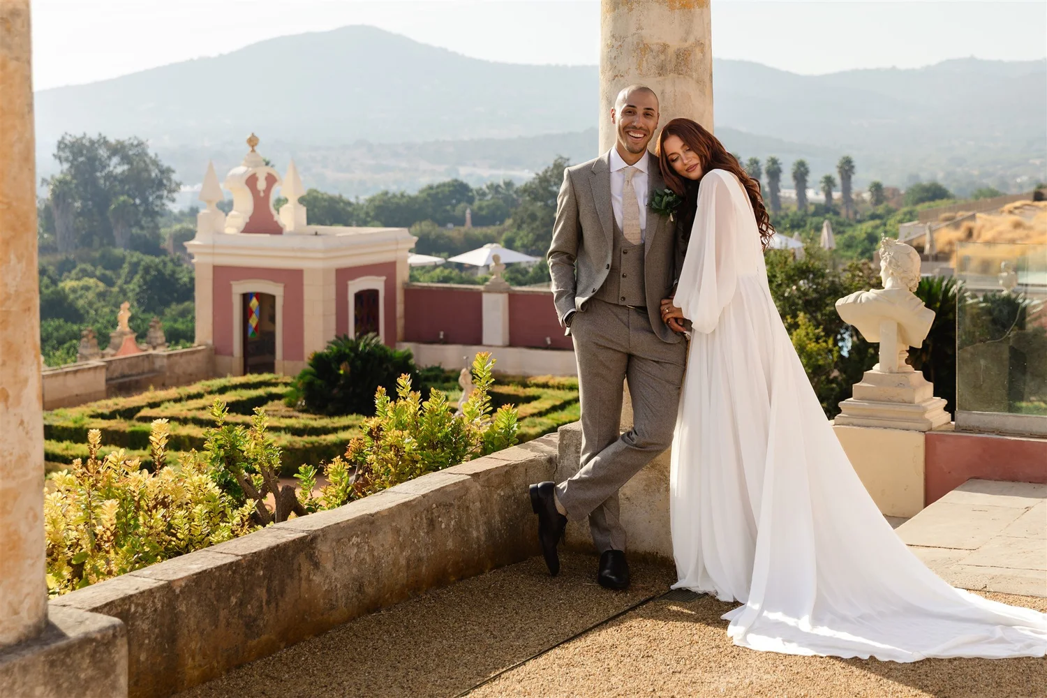 A smiling couple, dressed in wedding attire, stands on a terrace with decorative columns and sculptures, overlooking a lush landscape with mountains in the distance. The groom wears a gray suit, and the bride is in a white gown with long red hair. The setting appears to be an outdoor wedding venue with greenery, historic architecture, and scenic views.
