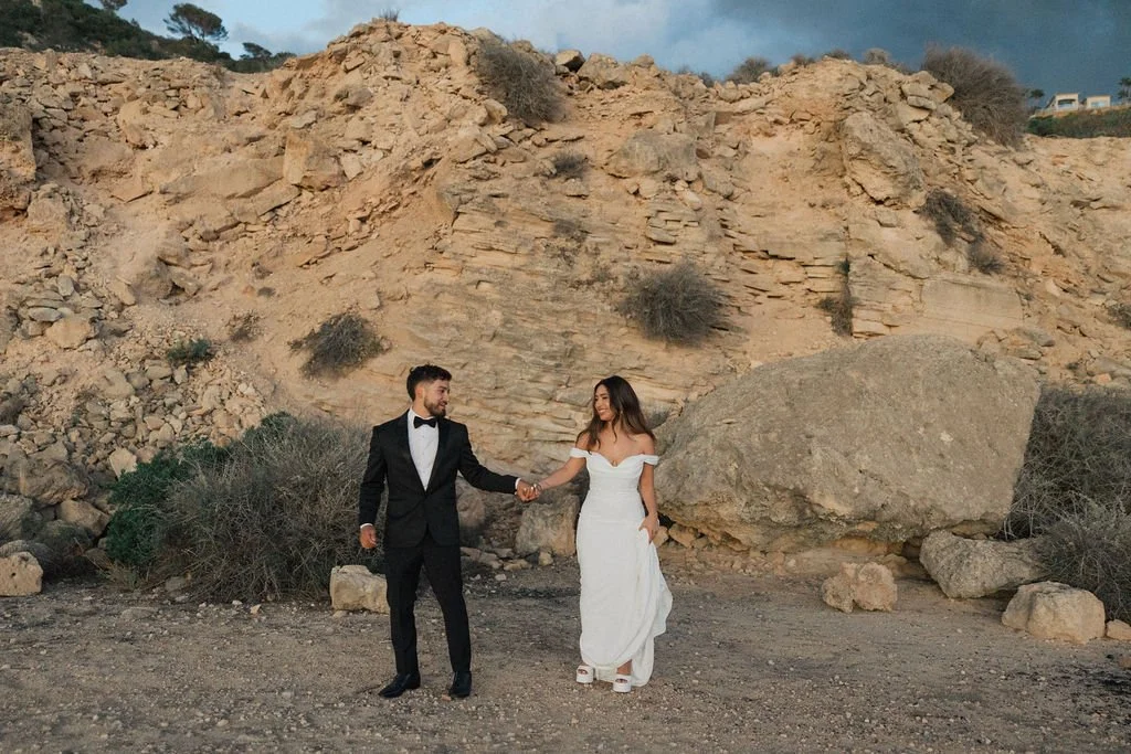 A couple dressed in wedding attire, holding hands, standing in front of a rocky desert landscape during the daytime.