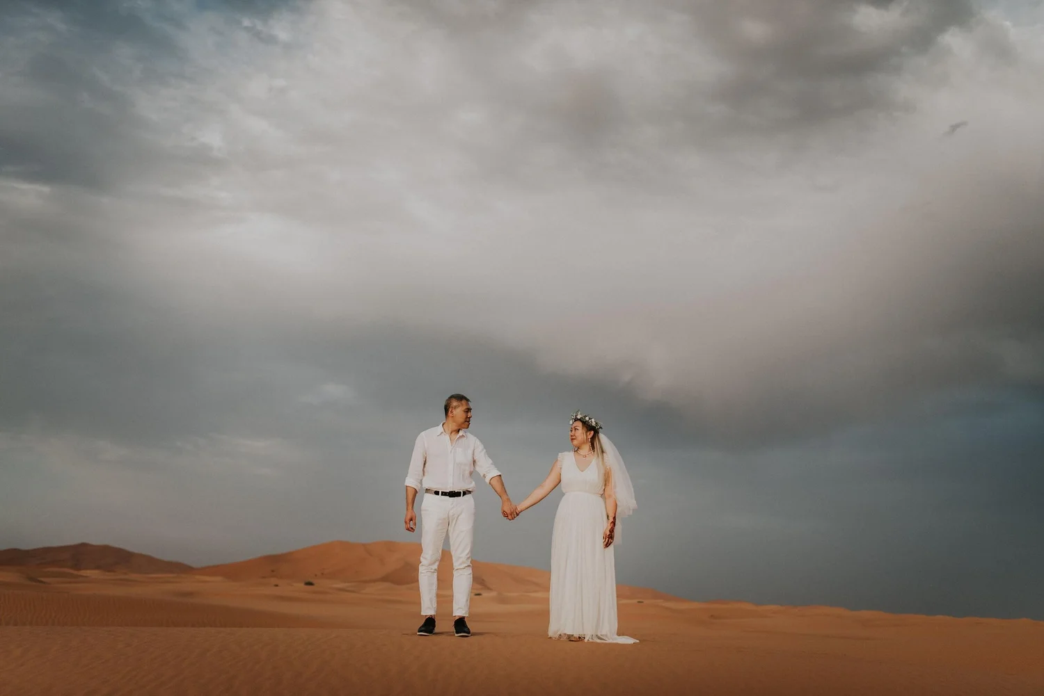 A couple dressed in wedding attire holding hands while walking in a desert landscape under a cloudy sky.