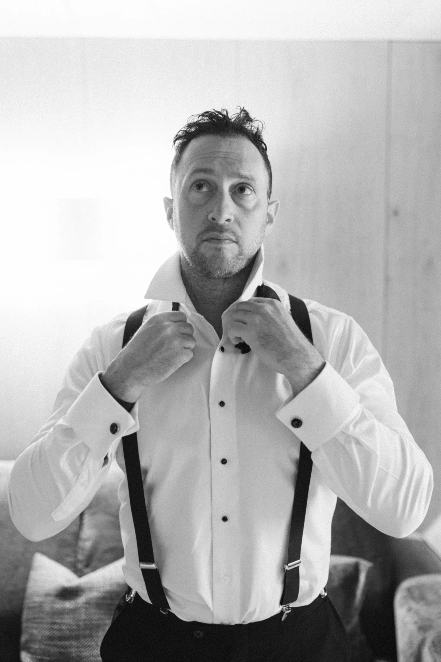 Man dressed in a white dress shirt, black suspenders, fixing his collar in front of a mirror.