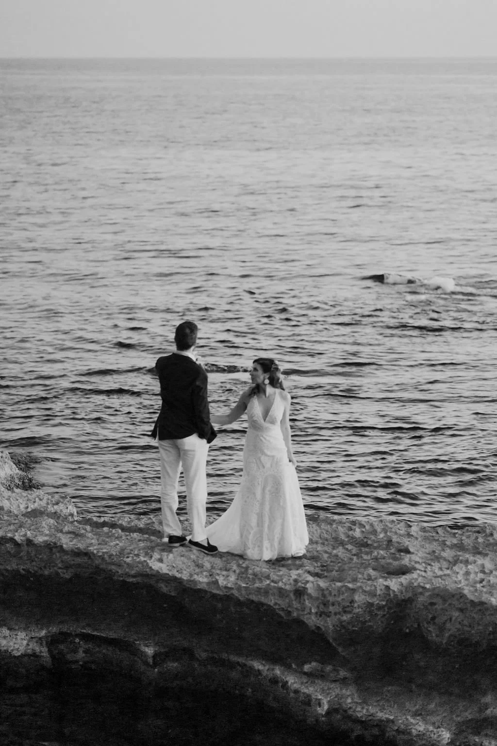 A black-and-white photo of a bride and groom holding hands on a rocky beach with the ocean in the background.