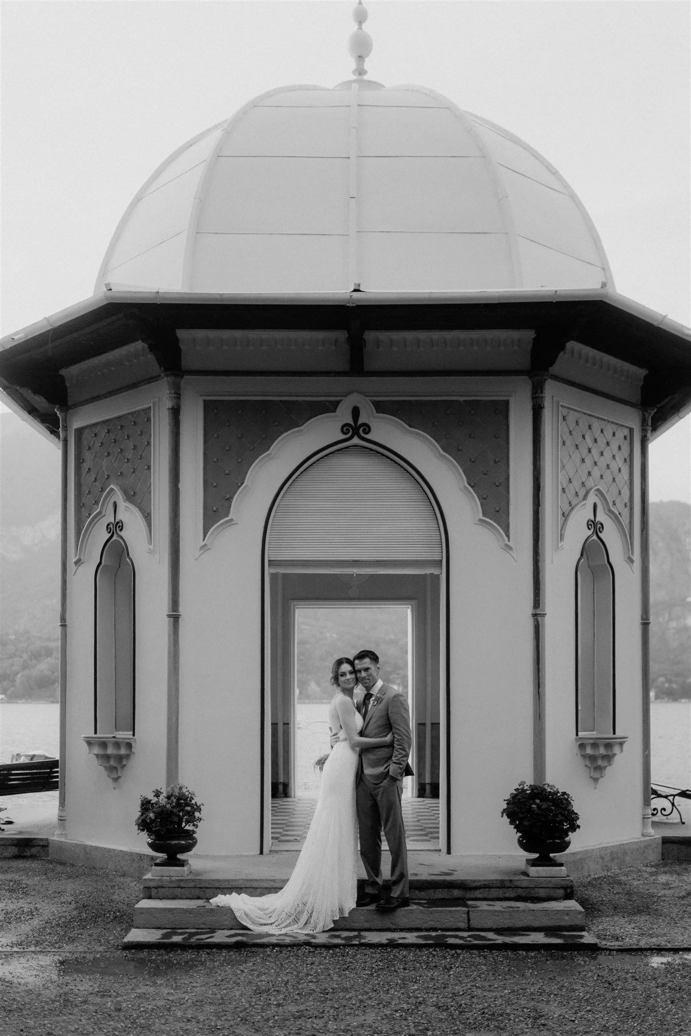 A bride and groom in wedding attire standing close together under a decorative pavilion by a body of water, with mountains in the background.