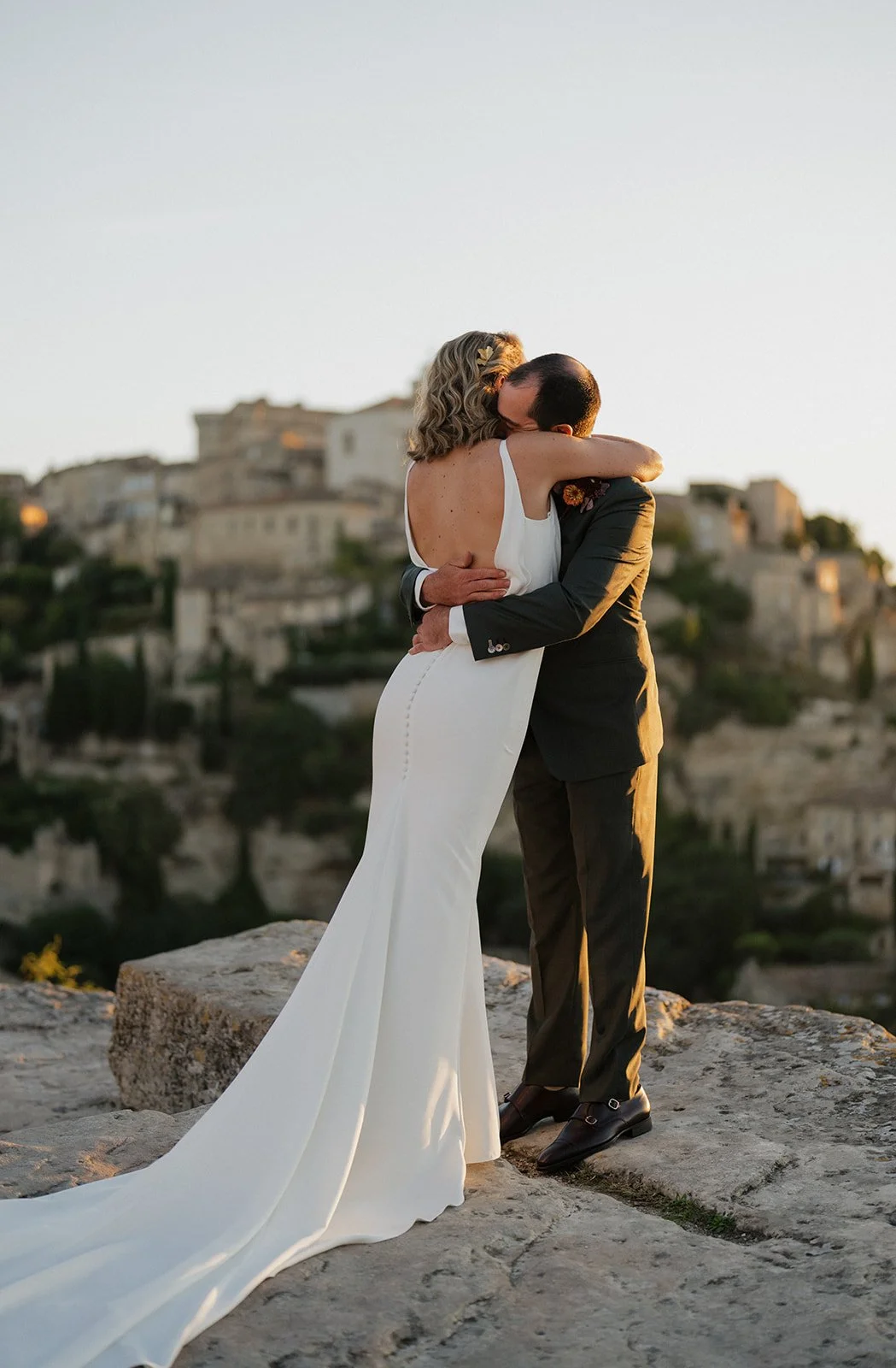 A newlywed couple embracing outdoors on a rocky ledge overlooking a historic hillside city during sunset.
