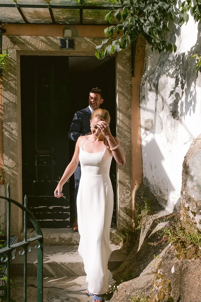 A woman in a white dress and bracelet walking out of a doorway, smiling and covering her face with her hand. A man in a suit is smiling in the background.