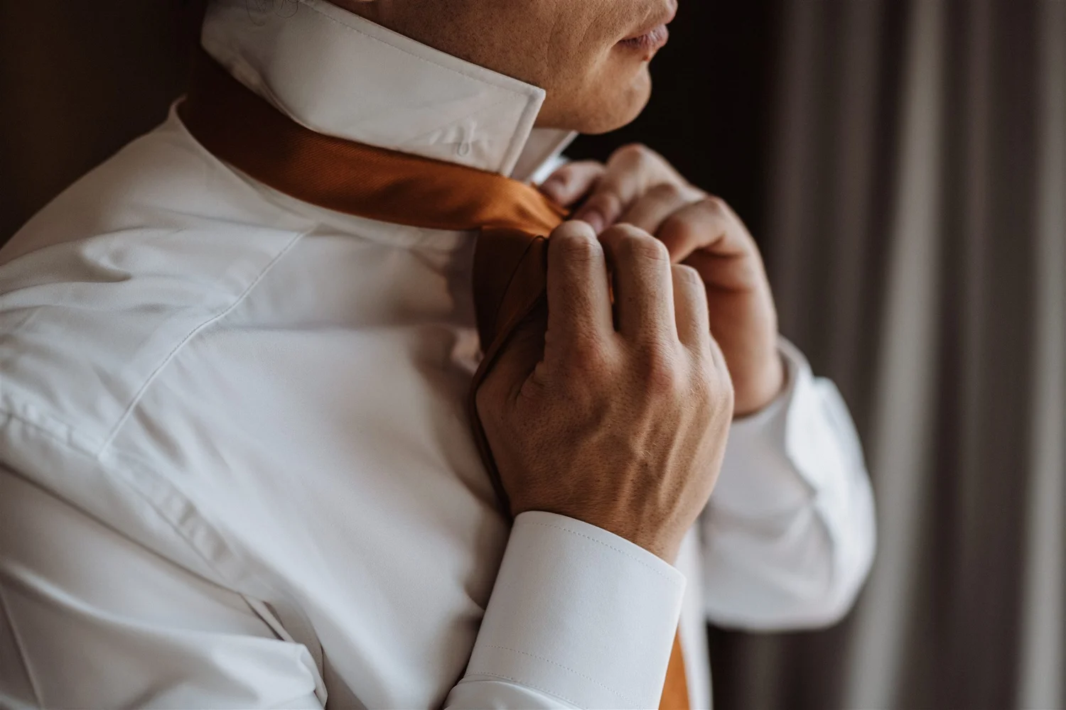A person is adjusting the collar of their white dress shirt, with a brown lining on the inside of the collar, standing near a curtain.