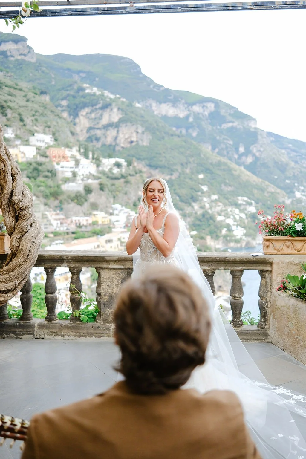 A bride in a wedding dress is smiling and clapping, standing on a terrace with potted plants and a stone railing, with a scenic mountainous landscape in the background. A person with brown hair, in a suit, is seen from behind in the foreground, watch