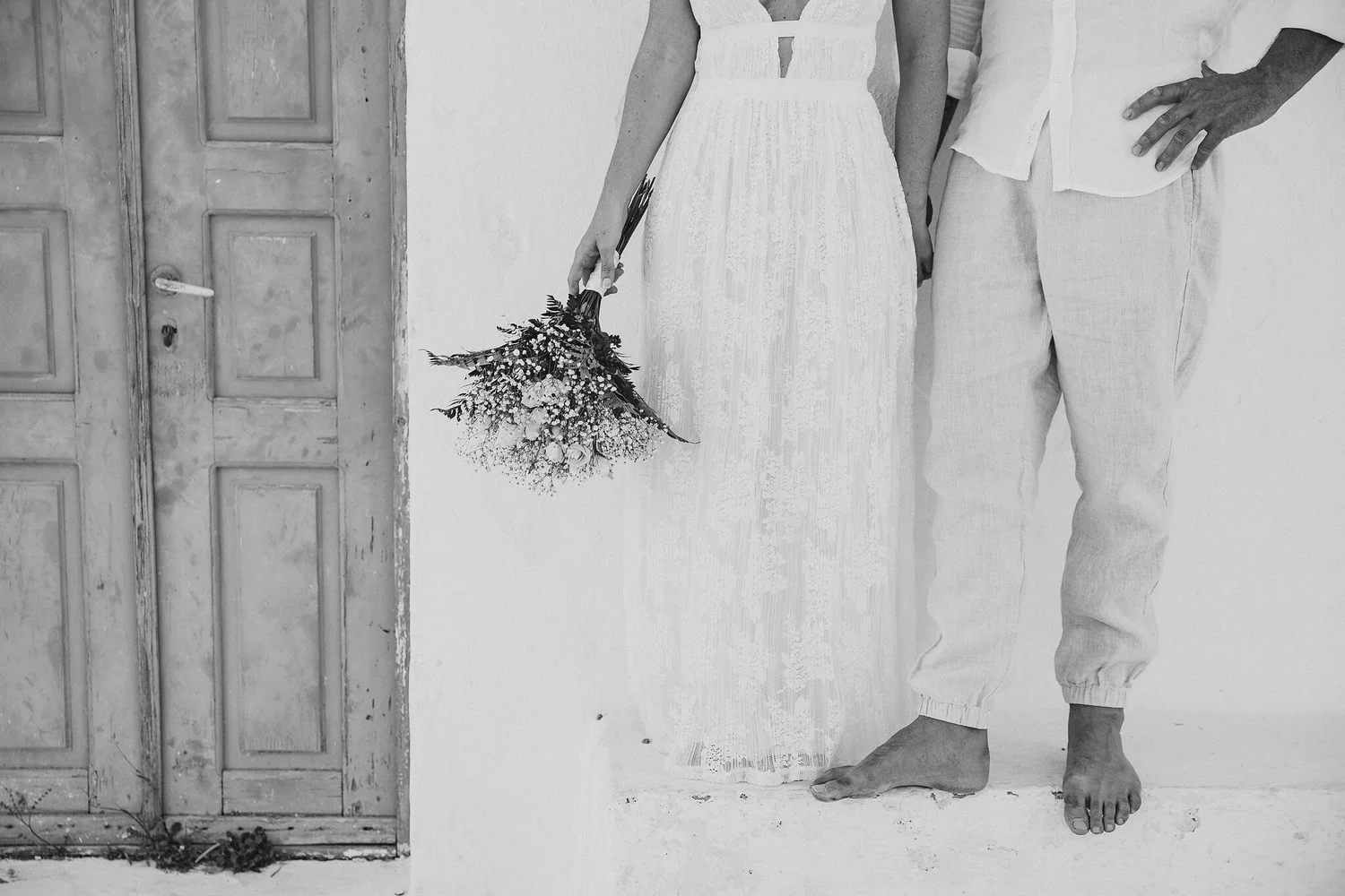Black and white photo of a woman in a long dress holding a bouquet of flowers standing next to a man in a white shirt and light-colored pants with one hand on his hip, standing barefoot in front of a wooden door with a handle and a keyhole.