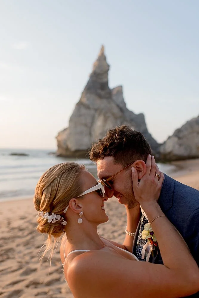A couple on a beach during sunset, embracing and smiling at each other with a large rock formation in the background.
