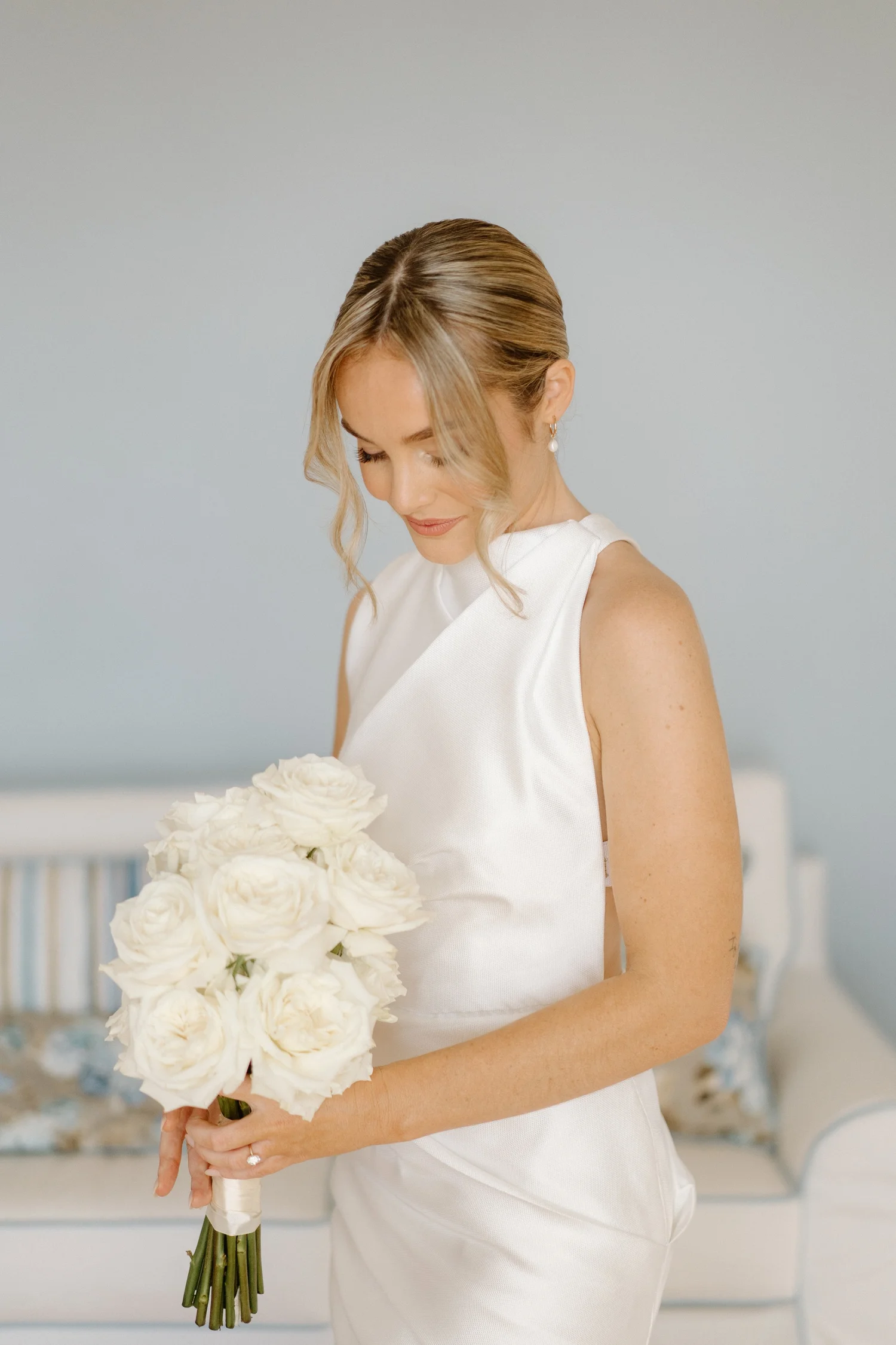 Woman in a white sleeveless dress holding a large bouquet of white roses, with an indoor background featuring chairs and a decorated wall.