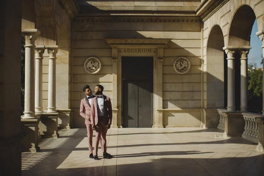 Two men in suits walk together in an outdoor area with stone columns and arches, in front of a building labeled 'AQUARIUM'.
