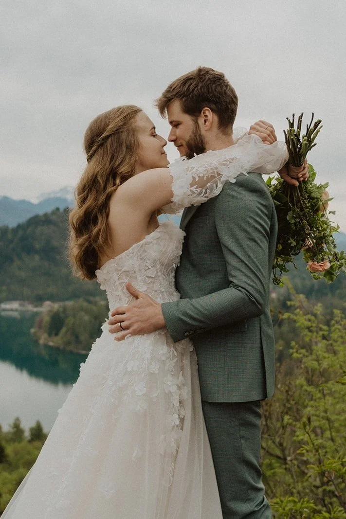 A bride and groom embrace outdoors with mountains and a lake in the background. The bride holds a bouquet of flowers.
