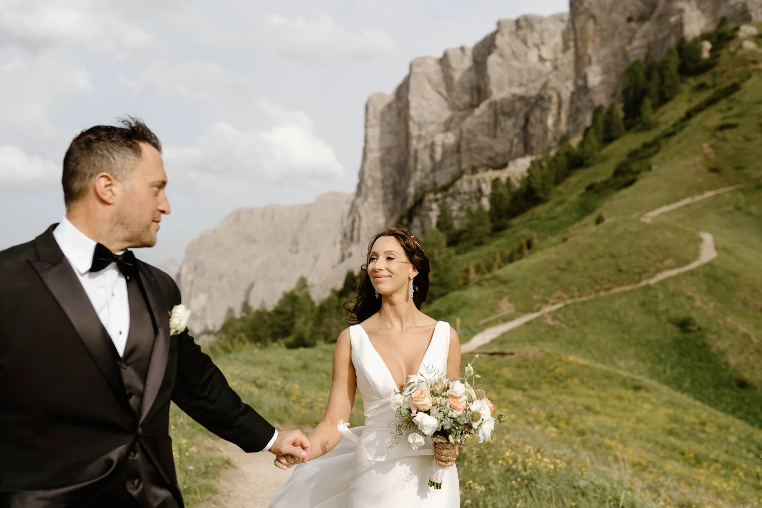 A bride and groom holding hands outdoors with a mountain and green hillside in the background. The bride is wearing a white wedding dress and holding a bouquet, and the groom is wearing a black tuxedo with a bow tie.