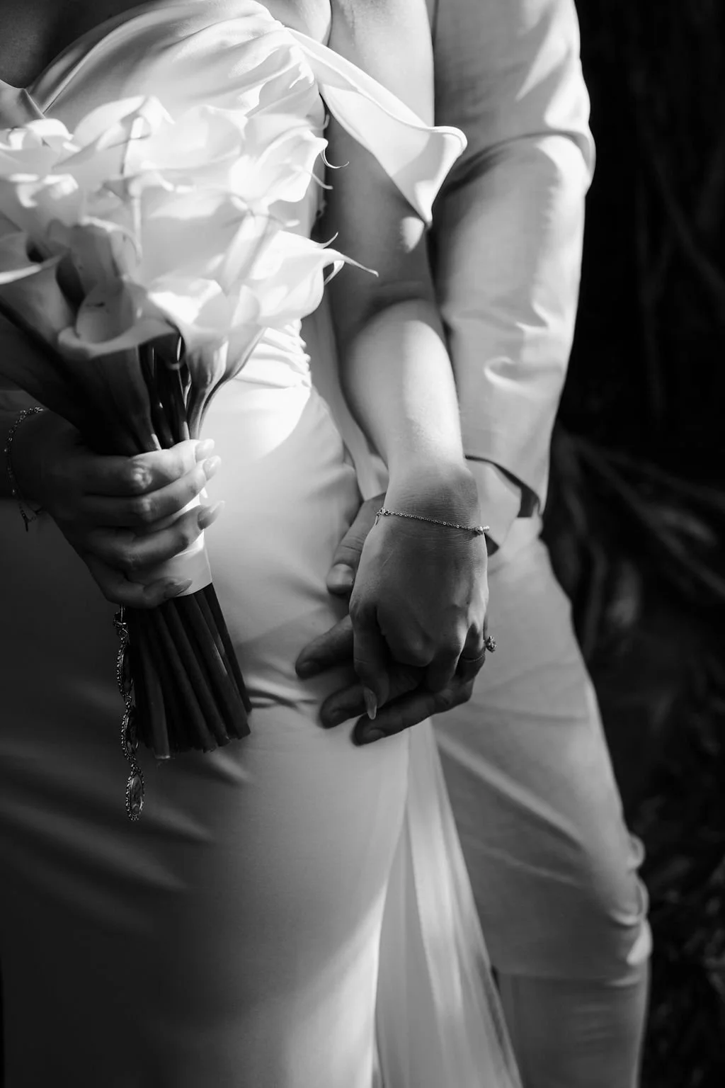 A black-and-white photo of a bride and groom holding hands during a wedding ceremony. The bride is holding a bouquet of flowers, and both are wearing wedding attire with the bride's dressed in a gown and the groom in a suit.