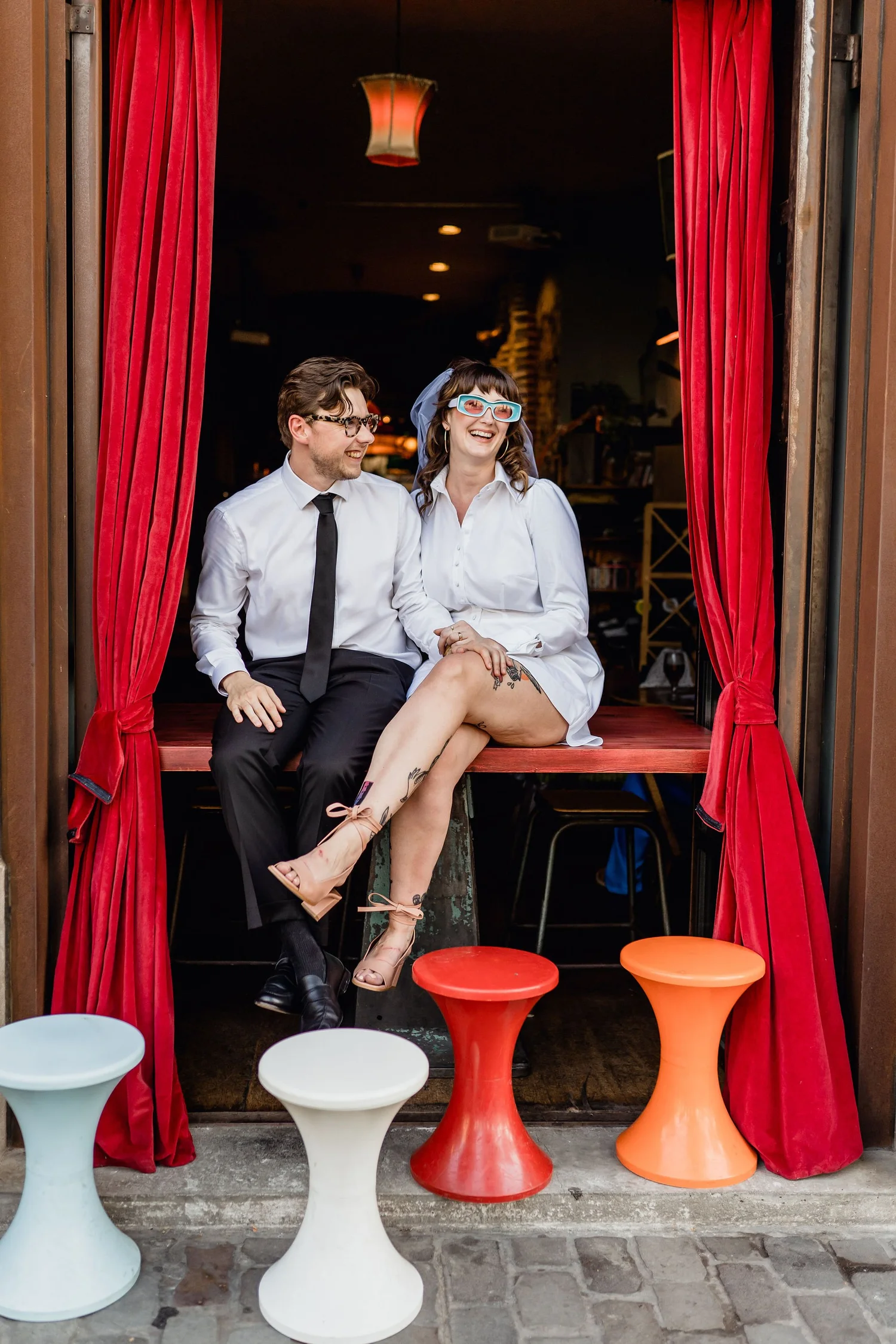 A man and woman sitting on a window ledge surrounded by red curtains, wearing glasses, with the woman also wearing a veil and light-up glasses, smiling and looking at each other.