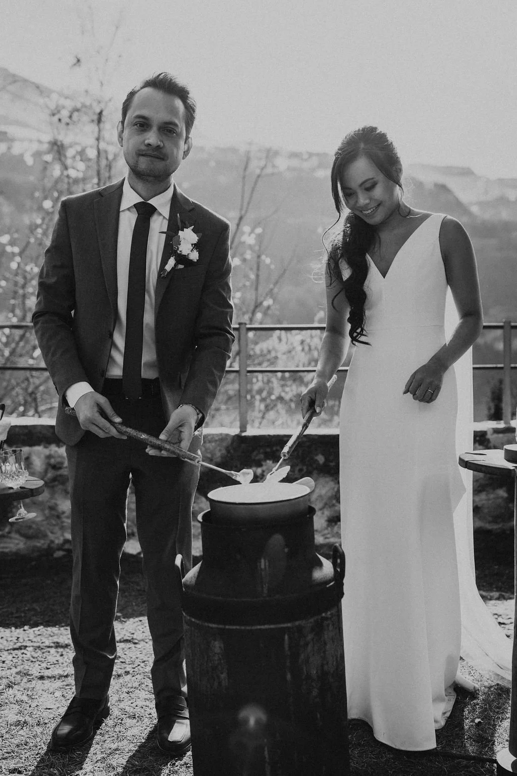 A black and white photo of a groom and bride cooking together outdoors near a mountain landscape. The man is wearing a suit with a boutonniere, and the woman is in a white dress. They are stirring a large pot, smiling and enjoying the moment.