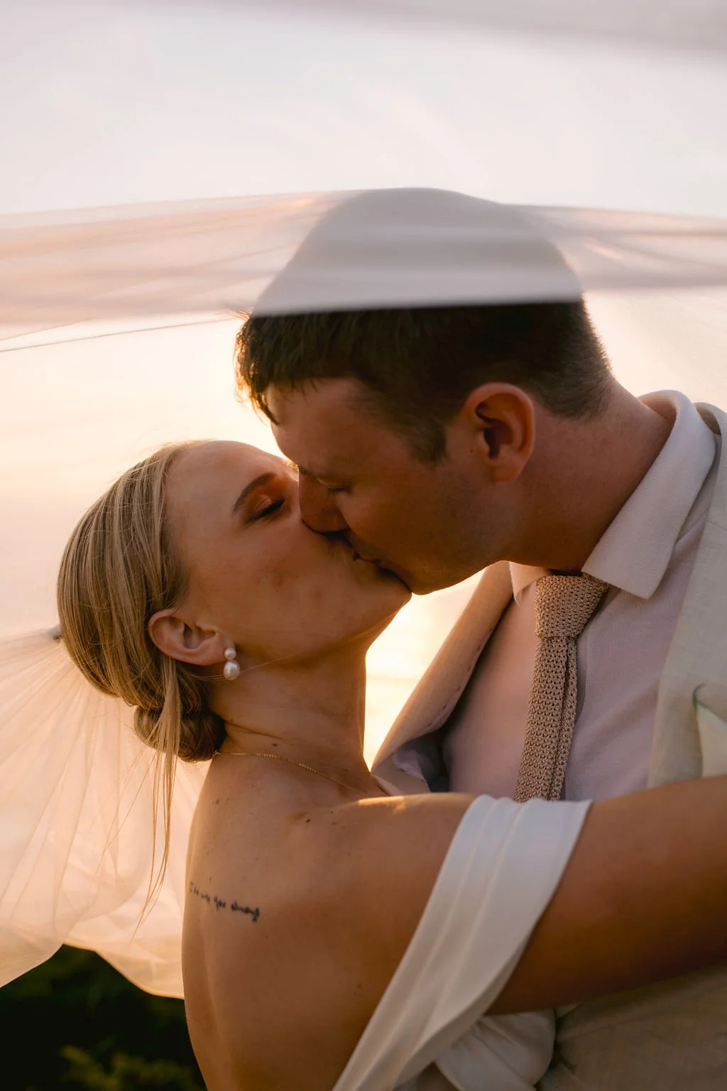 A bride and groom share a kiss under a wedding umbrella during sunset.