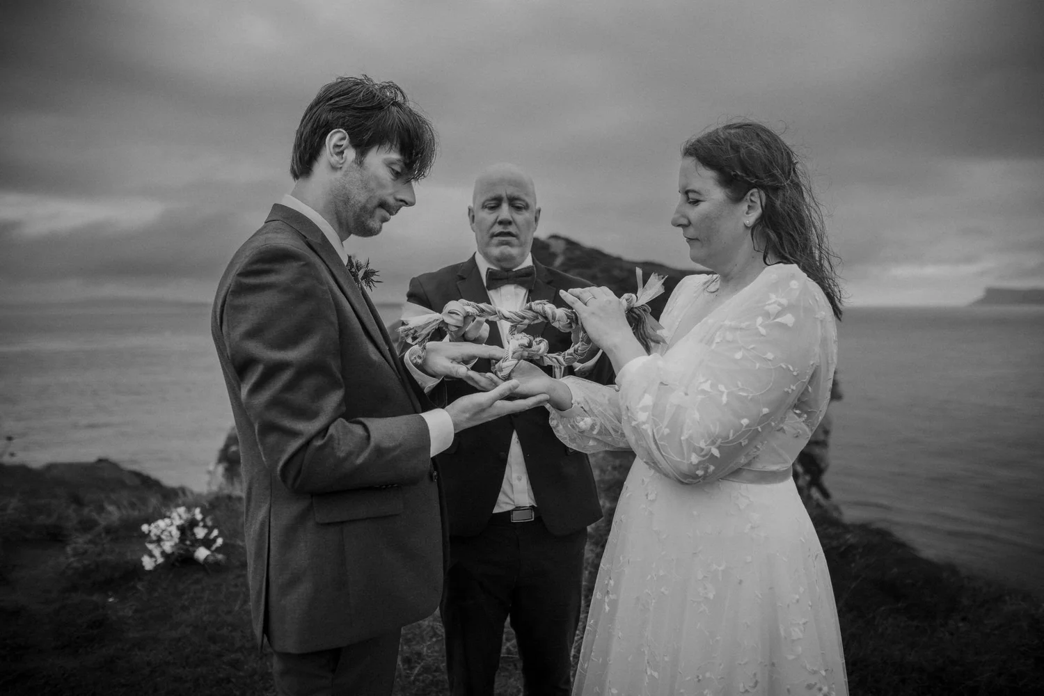 A black and white photo of a wedding ceremony on a beach, with a bride and groom holding hands and an officiant in the middle.