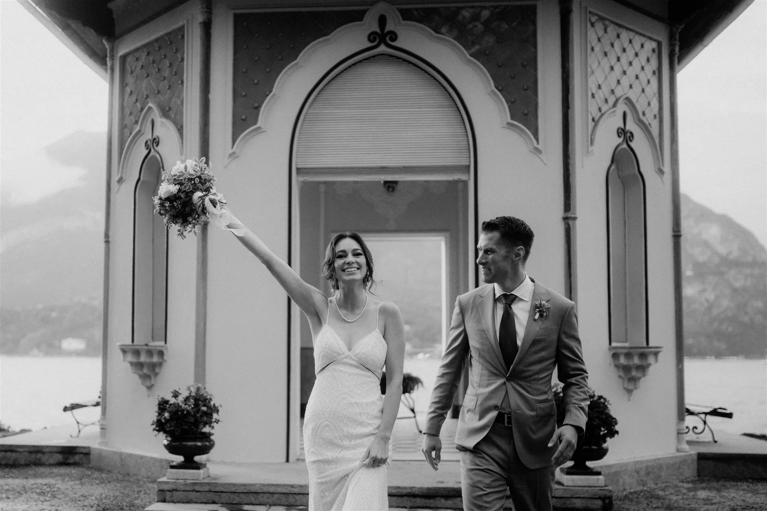 A joyful bride in a white wedding dress and a groom in a suit holding hands, walking out of a small outdoor chapel with decorative arches, while the bride raises a bouquet in celebration.