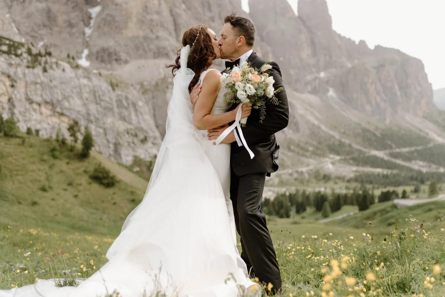 A bride and groom kissing outdoors in a green field with wildflowers, mountains in the background, the bride holding a bouquet of flowers, dressed in wedding attire.