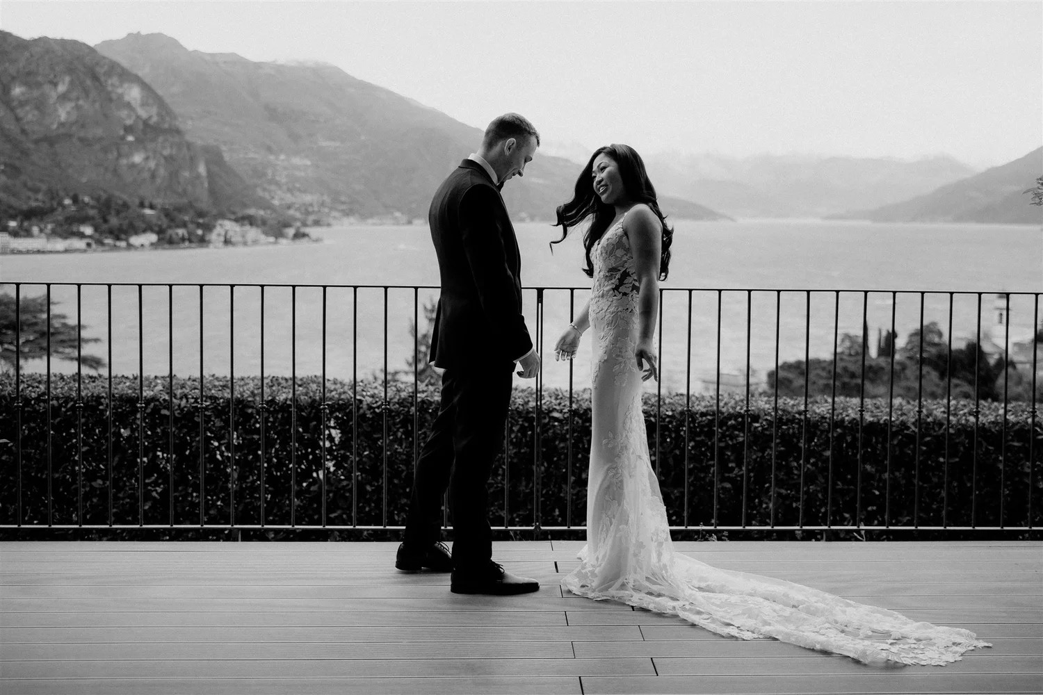 A bride and groom standing close to each other outdoors on a balcony, holding hands and smiling at each other, with mountains and a body of water in the background, in black and white.