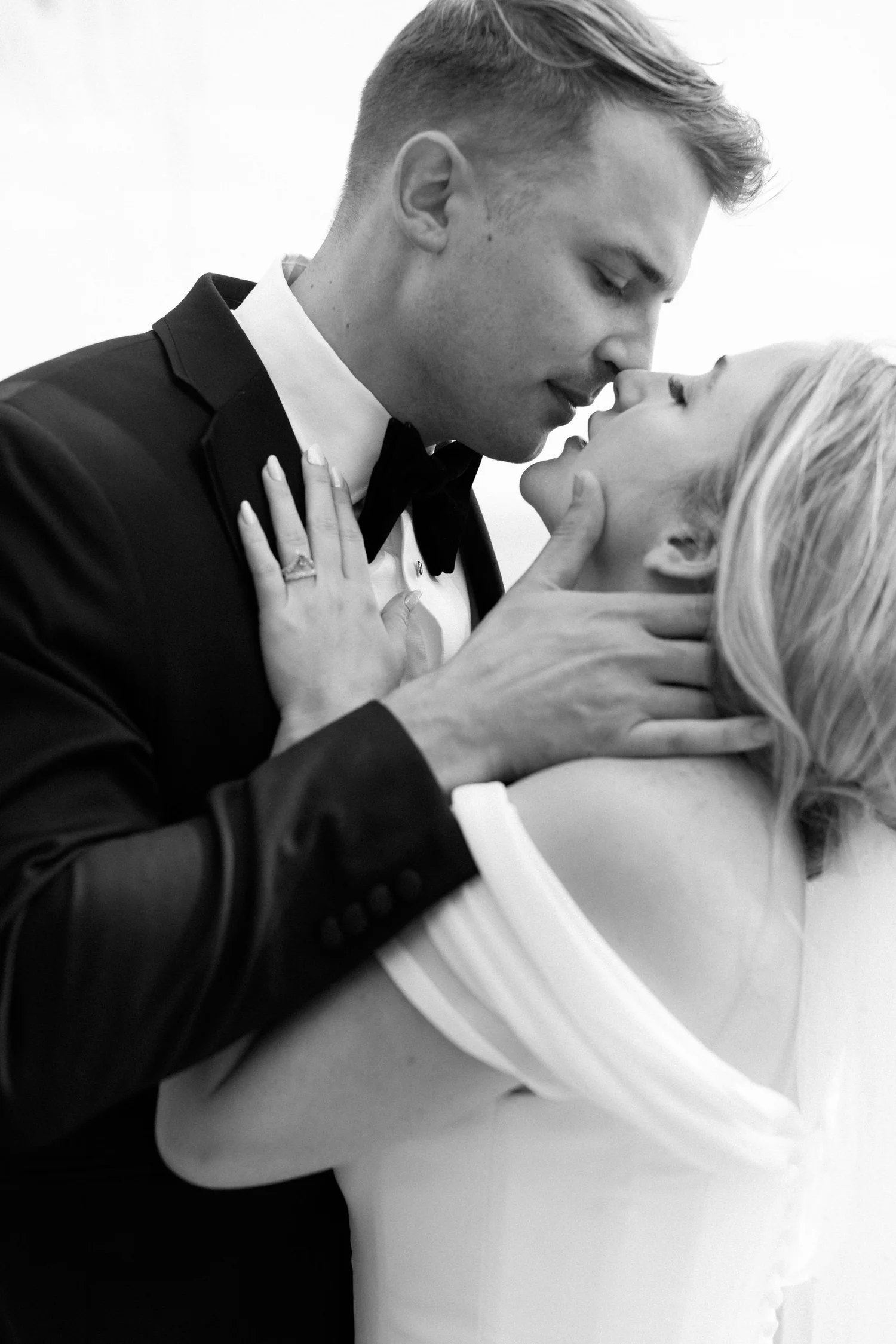 A black-and-white photo of a man and woman about to kiss, with the man's hand gently touching the woman's neck and her hand on his chest, wearing formal attire.