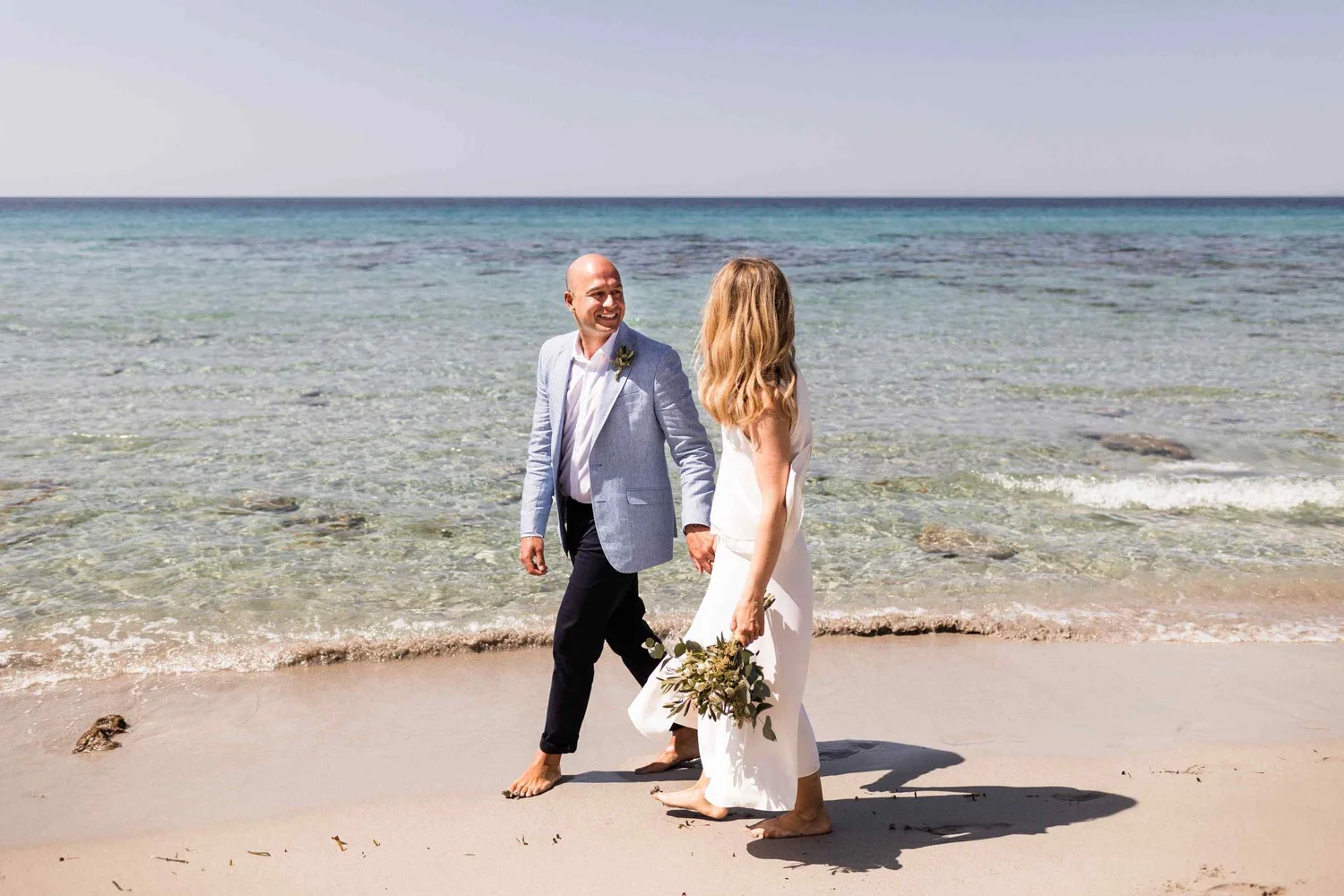 A couple, dressed in wedding attire, walks hand-in-hand along the beach with the ocean in the background. The bride wears a white dress and holds a bouquet of greenery, while the groom wears a light gray suit and has a boutonniere. Both are barefoot