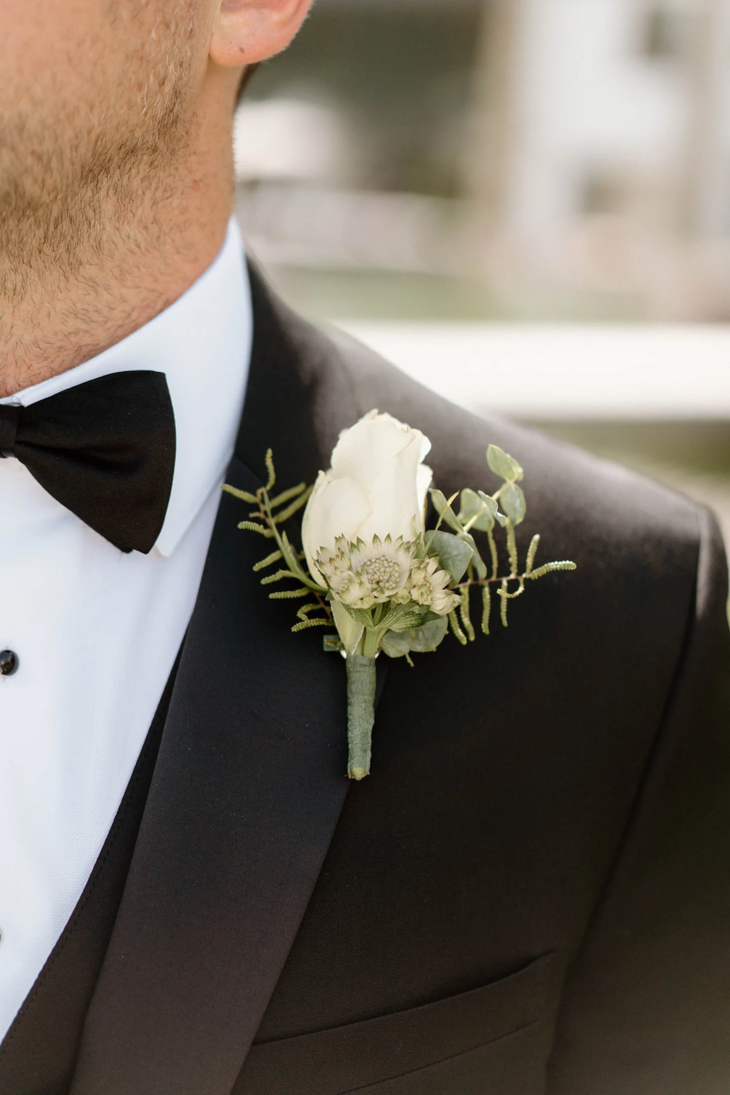 Close-up of a groom in a black tuxedo with a white shirt and black bow tie, wearing a white boutonniere with a cream-colored flower and greenery on the lapel.