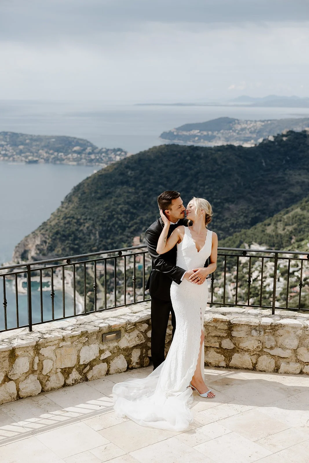 A newly married couple sharing a romantic kiss on a balcony overlooking a scenic coastal landscape with mountains, water, and a town below.