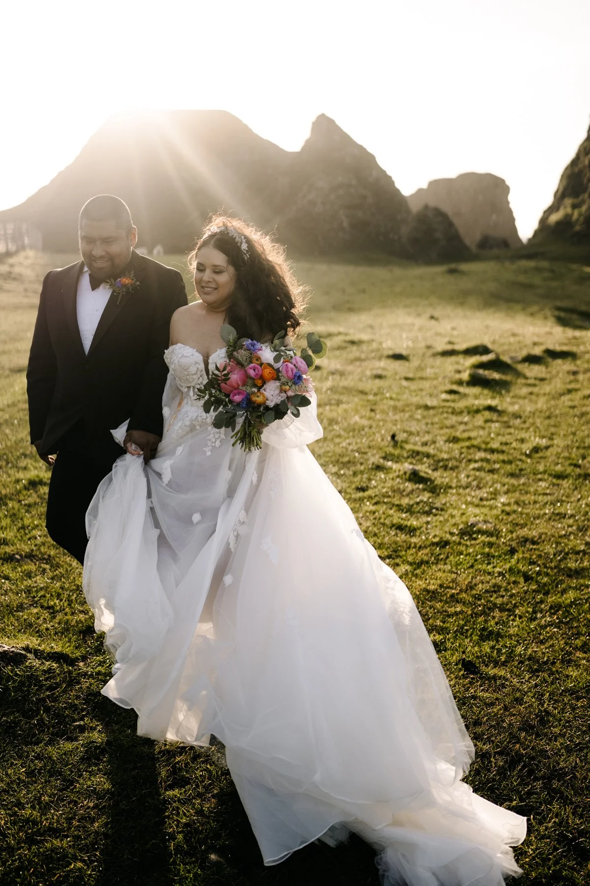 A bride and groom walking outdoors on a grassy field during sunset, with mountains in the background. The bride is wearing a white wedding gown and holding a colorful bouquet, and the groom is dressed in a black tuxedo with a boutonniere.