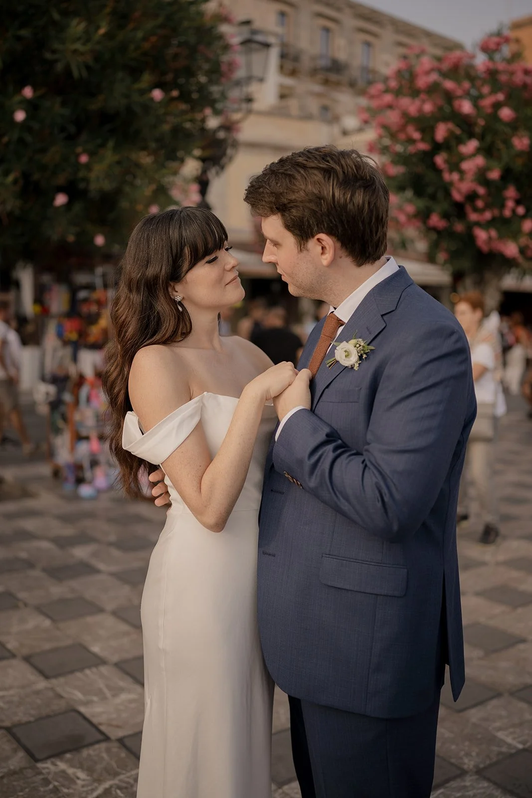 A bride and groom share a romantic dance outdoors in a city square, with pink blooming trees in the background during a wedding celebration.
