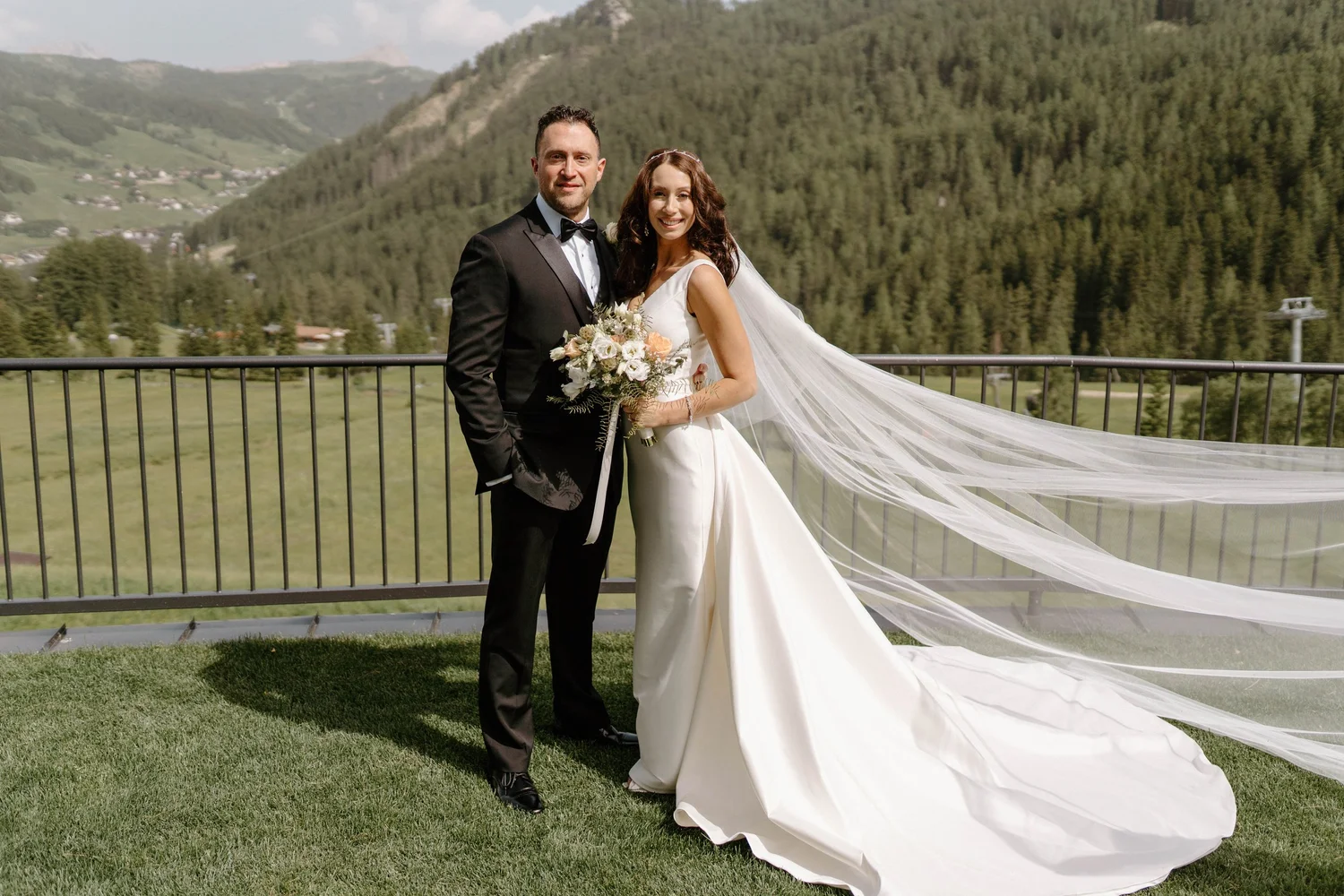 A bride and groom posing outdoors on a balcony with lush green mountains in the background. The bride is holding a bouquet and wearing a white wedding dress with a long veil. The groom is dressed in a black tuxedo. Both are smiling at the camera.