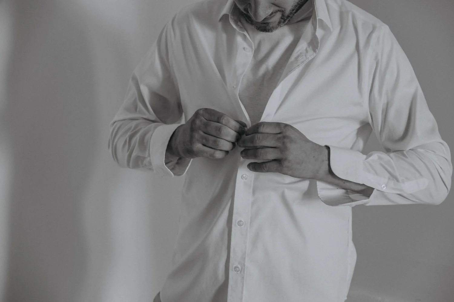 A man in a white shirt is buttoning his shirt, with his head tilted downward, against a plain background.