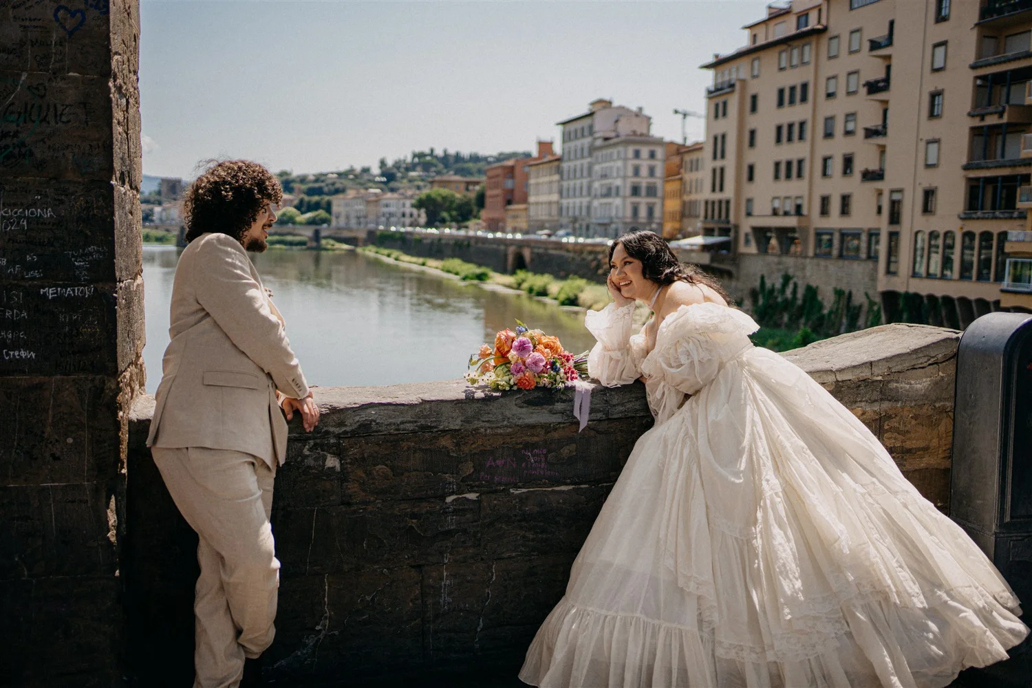 A man and a woman are standing near a river in an urban area, smiling and looking at each other. The woman is wearing a white wedding dress and holding a bouquet, suggesting a wedding or celebration. The man is dressed in a light-colored suit. There are tall buildings and a distant hill in the background.