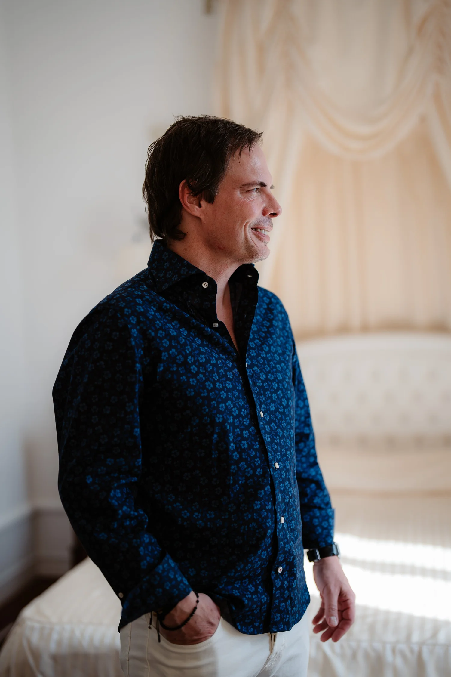 A man with dark hair and light skin is standing in a room, wearing a dark patterned shirt and light-colored pants. He is smiling and has his hands in his pockets, with a watch and wristbands visible on his left wrist. The background features a cream-