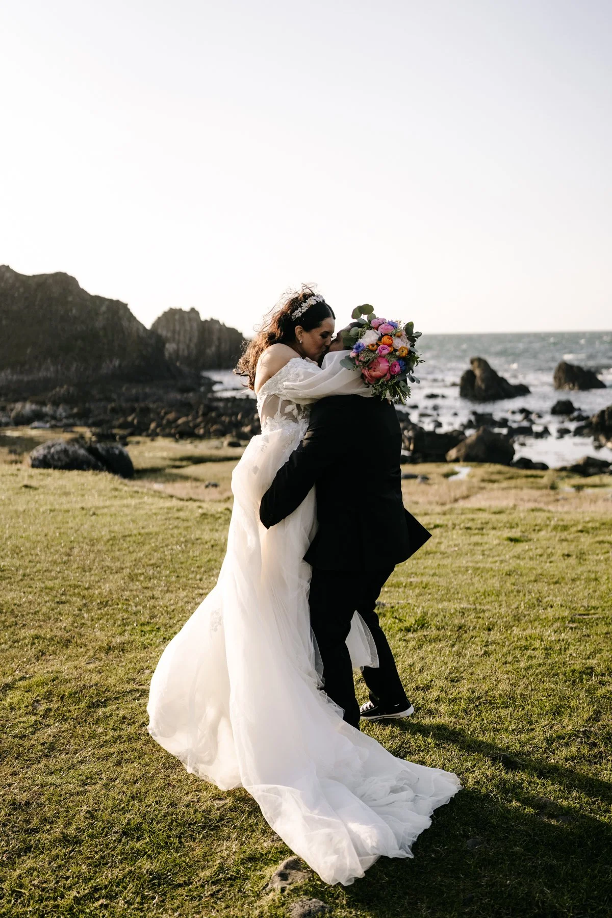 A bride and groom embrace on a grassy field near the ocean, with rocks and cliffs in the background, during sunset.