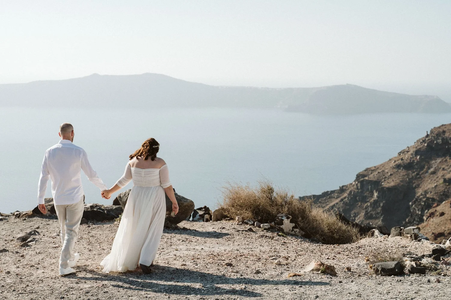 A couple holding hands and walking along a rocky beach with mountains and water in the background.