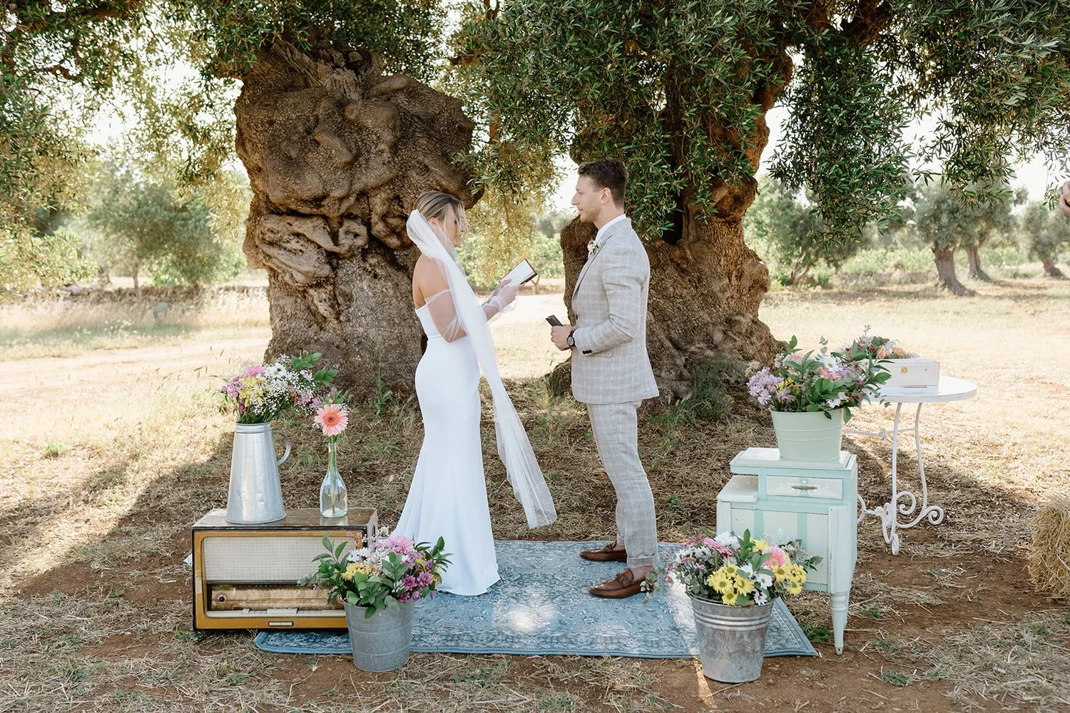 A wedding ceremony taking place outdoors under a large tree, with a bride and groom standing facing each other, exchanging vows. The bride is in a white dress with a veil, and the groom in a light gray plaid suit. There are floral arrangements on the