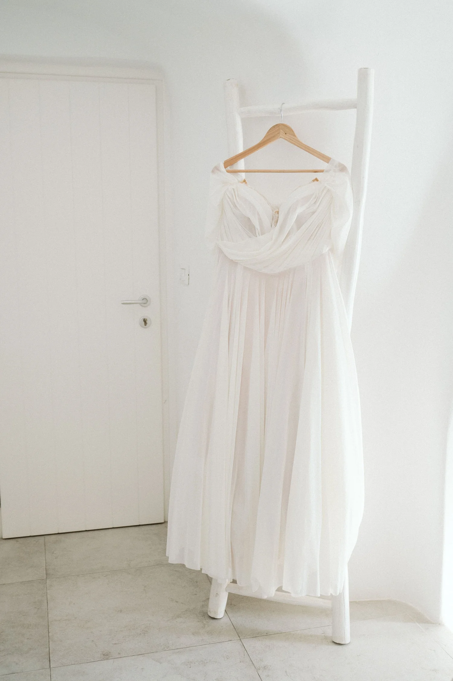 A white wedding dress hanging on a wooden hanger on a white chair in front of a white wall.