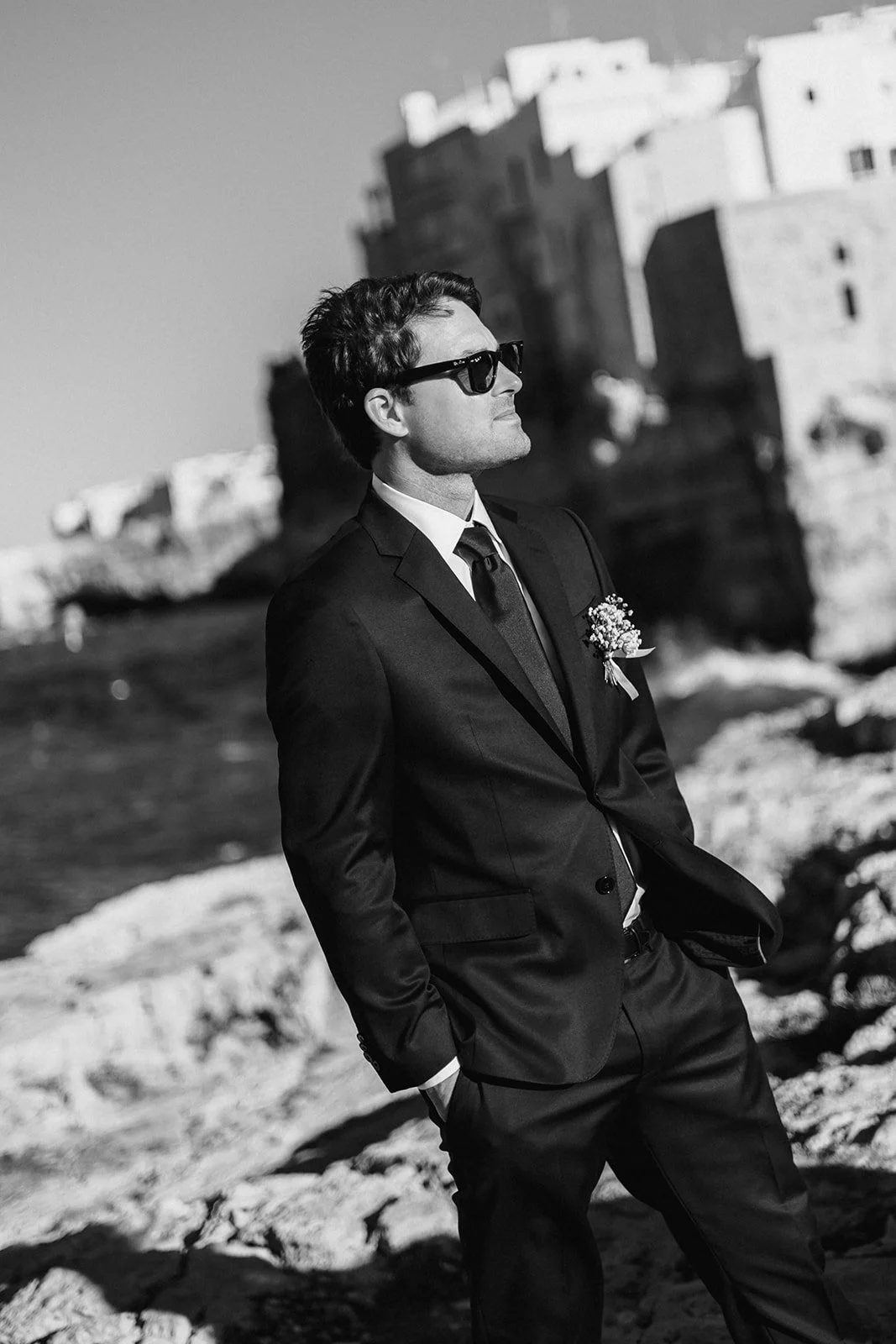 A man in a suit and sunglasses standing outdoors near the water, with an old stone castle or fortress in the background.