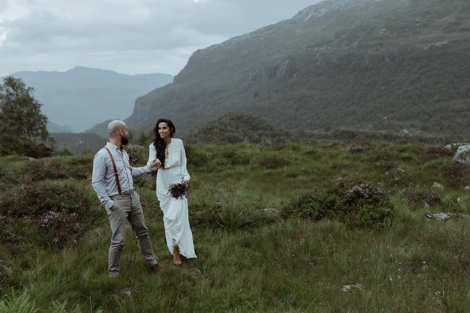 A couple, a man with a bald head and beard in a light shirt with suspenders and a woman in a white dress with long dark hair, standing hand in hand in a grassy field with mountains in the background during overcast weather.