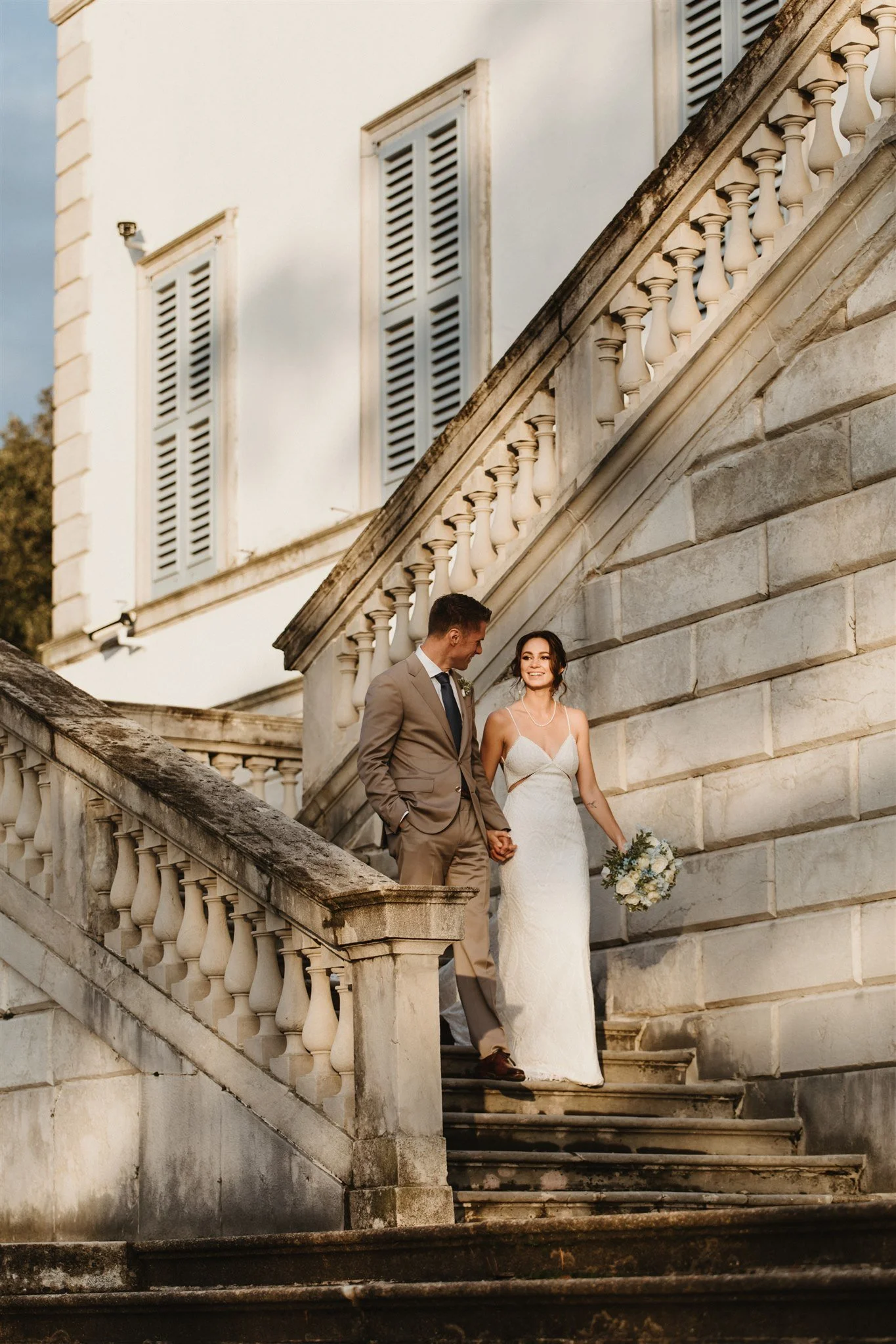 A newlywed couple stands on an outdoor stone staircase, holding hands, with the bride holding a bouquet of white and blue flowers, celebrating their wedding in front of an elegant building with closed shutter windows.