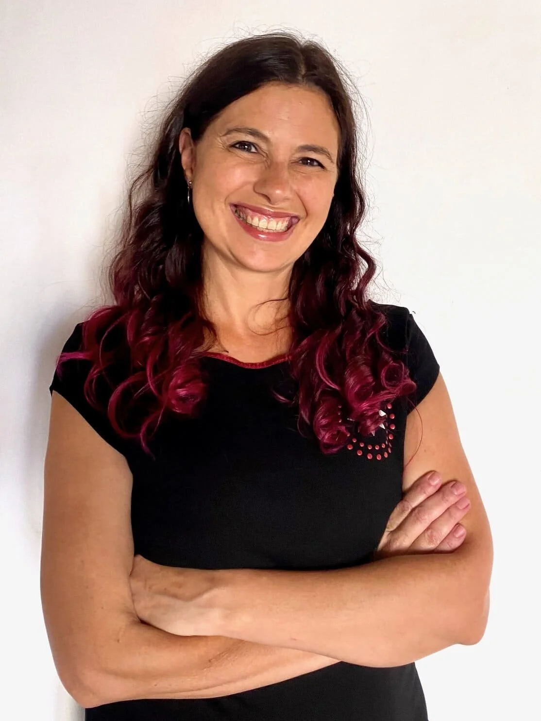 A woman with dark hair and pink curled ends, smiling with arms crossed, wearing a black shirt with red accents, standing against a plain white background.