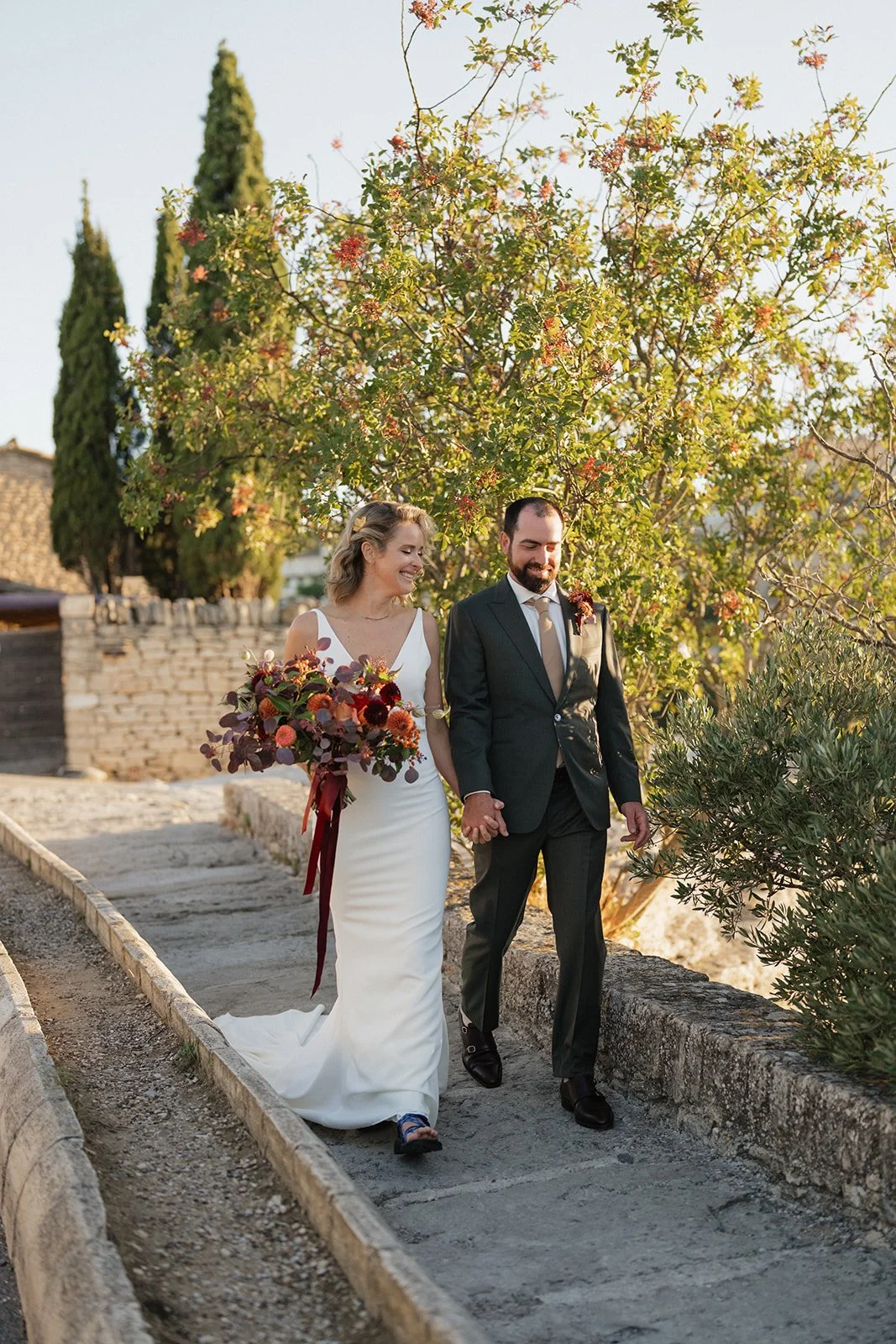 A bride and groom walking hand in hand outdoors during a wedding, smiling and holding hands, with greenery and a tree in the background.